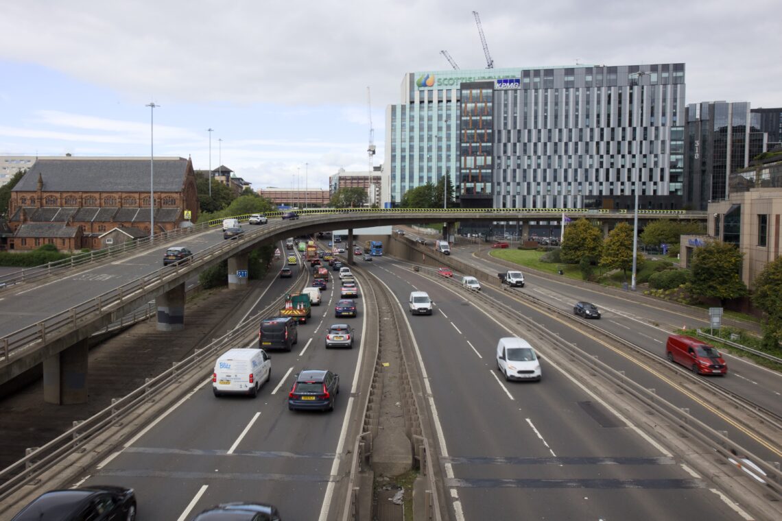 A busy multi-lane dual carriageway in a city with cars and vans travelling in both directions. A flyover crosses above, and a modern glass office block stands to the right. To the left is an old brick church. The sky is partly cloudy, and there are some cranes in the background.