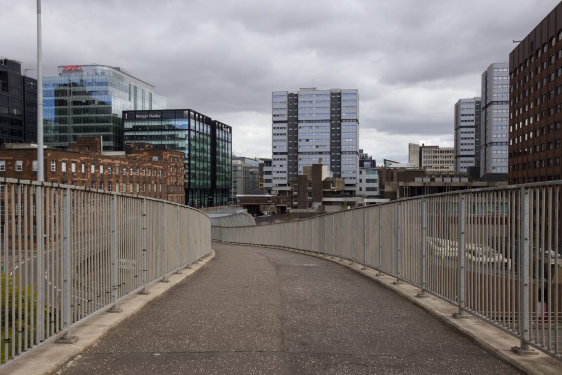 A pedestrian footbridge with metal railings curves gently through an urban area lined with modern glass buildings and older brick structures under a cloudy, overcast sky. Tall office blocks dominate the background.