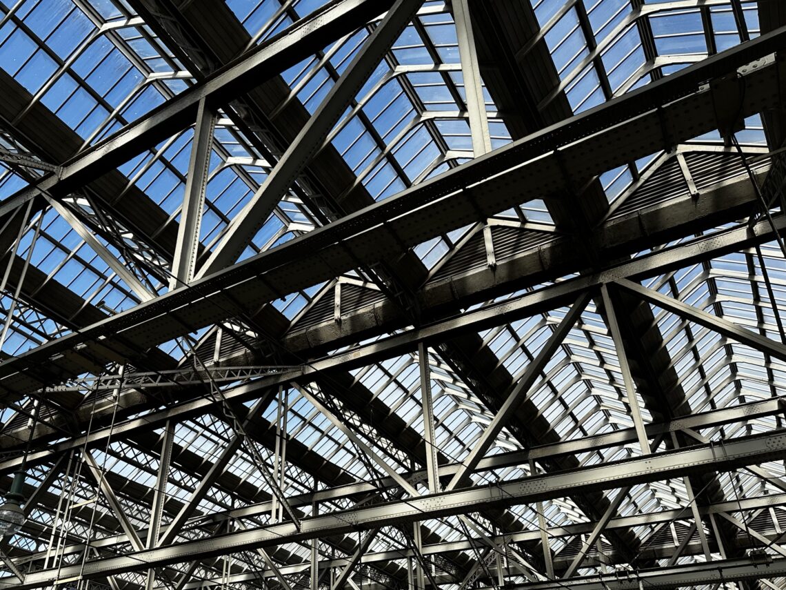 Steel beams criss-cross under a large glass roof, forming a geometric pattern. The blue sky is visible through the glass panels, and sunlight casts shadows across the industrial metal framework, suggesting a spacious, open interior.