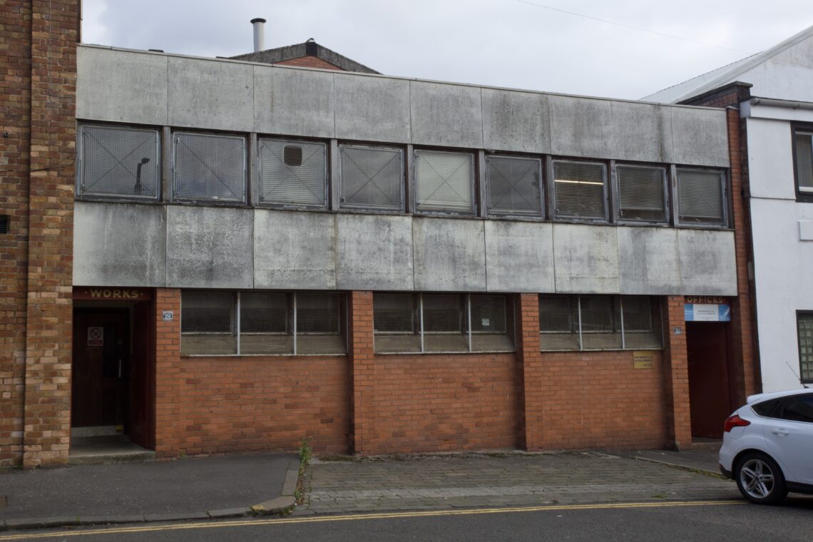 A two-storey industrial building with red brick on the ground floor and weathered grey concrete above. The windows are dirty and barred. There’s a door on the left and a parked white car partially visible on the right side of the image.