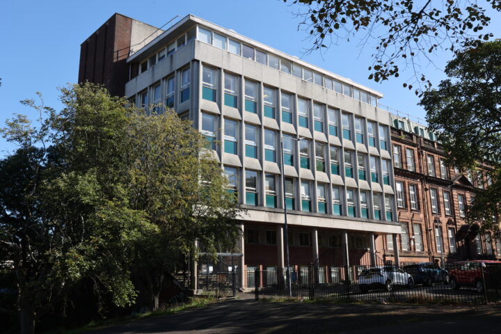 A five-storey modern building with large windows and a concrete façade stands next to older red-brick buildings. Trees partially obscure the lower floors, and parked cars are visible along the street in bright sunlight.