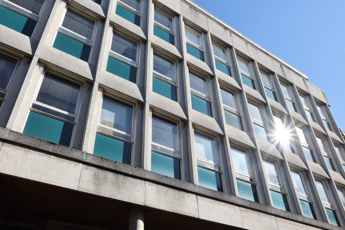 A concrete building with rows of large windows reflects bright sunlight. The photo is taken from a low angle, emphasising the modern, rectangular design. The sky is clear and blue, with the sun creating a starburst effect on the glass.