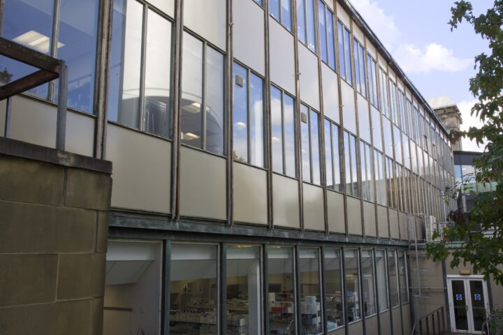 A modern building with large glass windows reflecting the sky and trees. The ground floor reveals a laboratory inside with equipment and shelves, while the upper floors show reflections and some desks with lights on. A tree is at the right edge.
