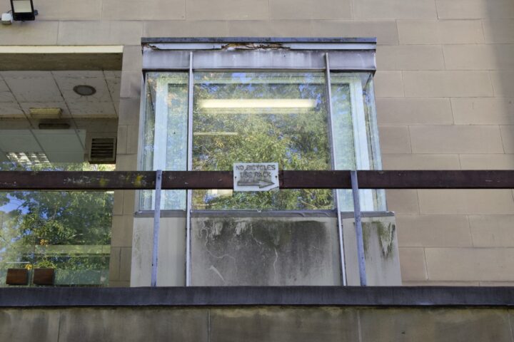 A weathered window with streaked glass and a rusted metal frame on a beige concrete wall. A sign reading NO BICYCLES TO BE PARKED is attached to a horizontal bar in front of the window. Green trees are faintly reflected in the glass.