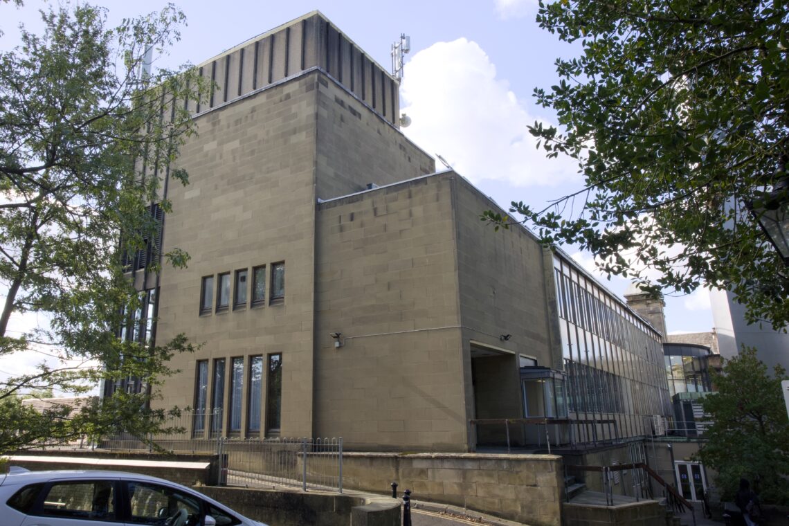 A large, modern building with beige stone walls and tall windows, including a section with vertical glass panels. Trees partially frame the building, and a parked car is visible in the lower left corner. The sky is partly cloudy.