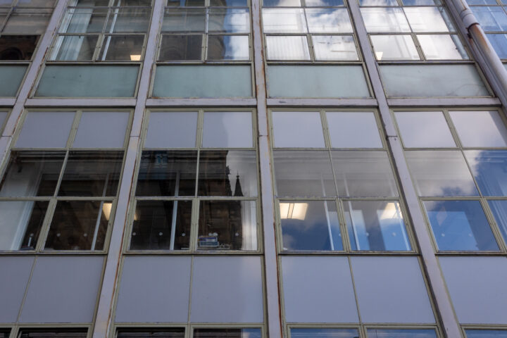 A close-up view of a modern building’s exterior, featuring large glass windows in a grid pattern. Some windows reflect the sky and clouds, whilst others reveal fluorescent lights and faint interiors. The window frames are metal and slightly weathered.