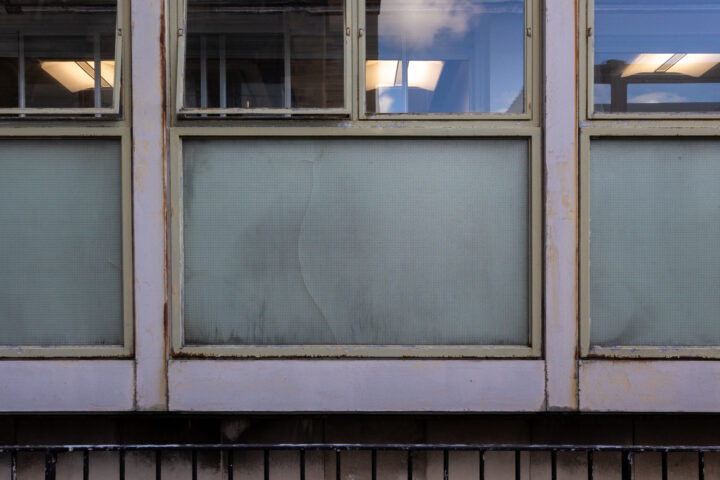 A close-up of three large, weathered windows in a beige building. The upper window panes are clear, showing lights and reflections, whilst the lower panes are frosted and stained, with visible dirt, cracks, and wear. A metal railing is at the bottom.