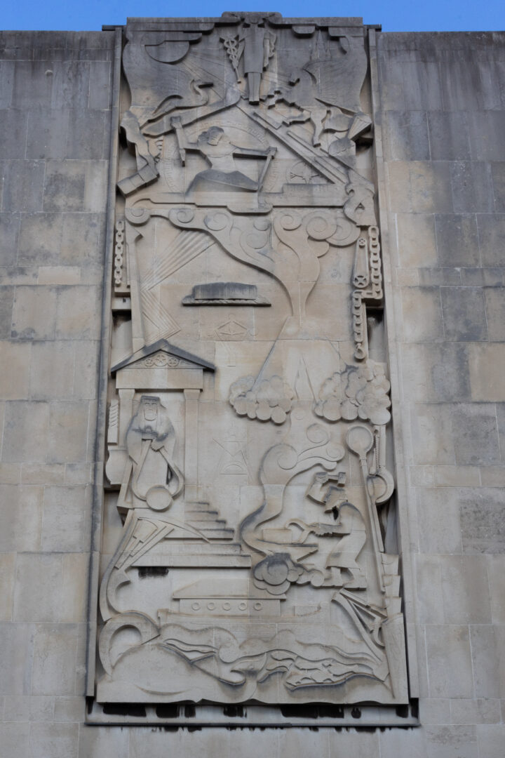 A tall stone relief sculpture on a building depicts abstract and figurative scenes: workers on rooftops, a staircase with a figure holding an object, trees, seated figures, clouds, smoke, and industrial elements, all richly detailed in geometric, stylised forms.