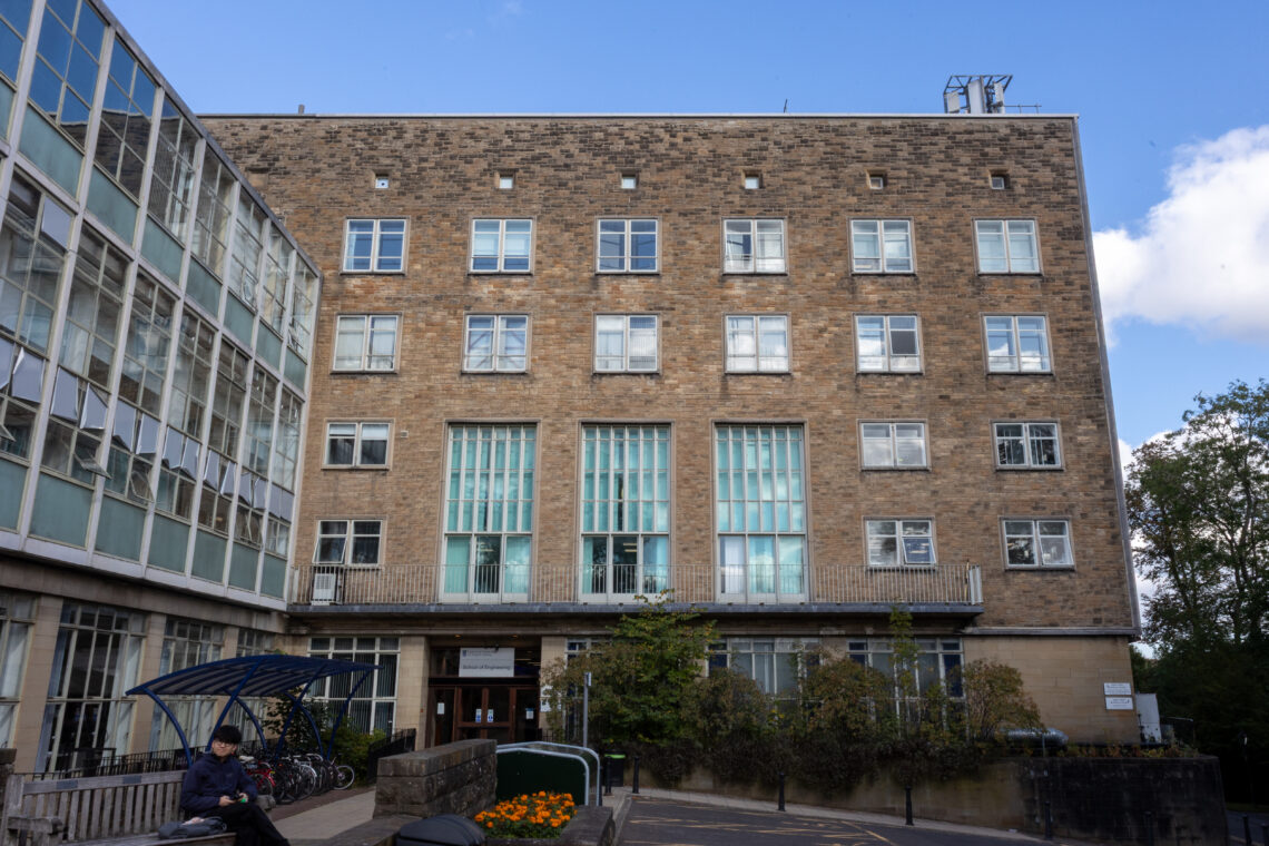 A multi-storey brick building with many windows, three tall blue-tinted window sections in the centre, and a glass-sided extension on the left. A person sits on a bench near flowerbeds, and trees frame the right side. The sky is partly cloudy.