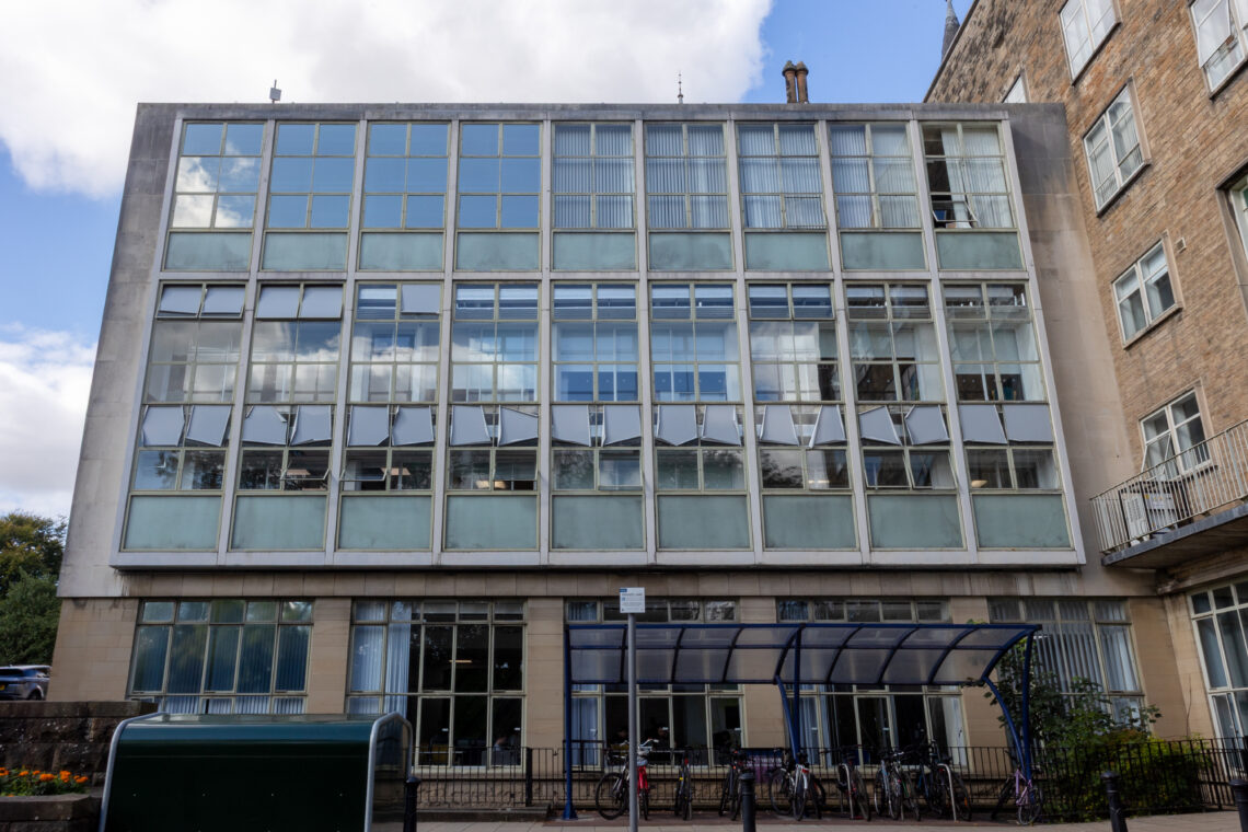 A modern building with large grid-like glass windows, four storeys tall. There is a blue-tinted cycle shelter with bicycles parked outside. Part of an older stone building is visible on the right. The sky is partly cloudy above.