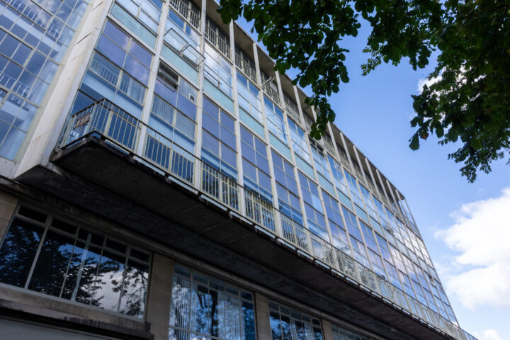 Low-angle view of a modern building with large blue-tinted glass windows and concrete balconies. Tree branches with green leaves frame the top of the image, and reflections of the sky and foliage are visible in the windows. Sky is partly cloudy.