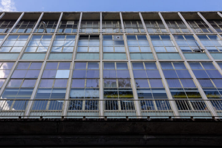 A low-angle view of a modern building facade with large grid-like glass windows, some panels are tinted or open. The building has metal railings and a concrete base, with a blue sky and clouds reflected in the glass.