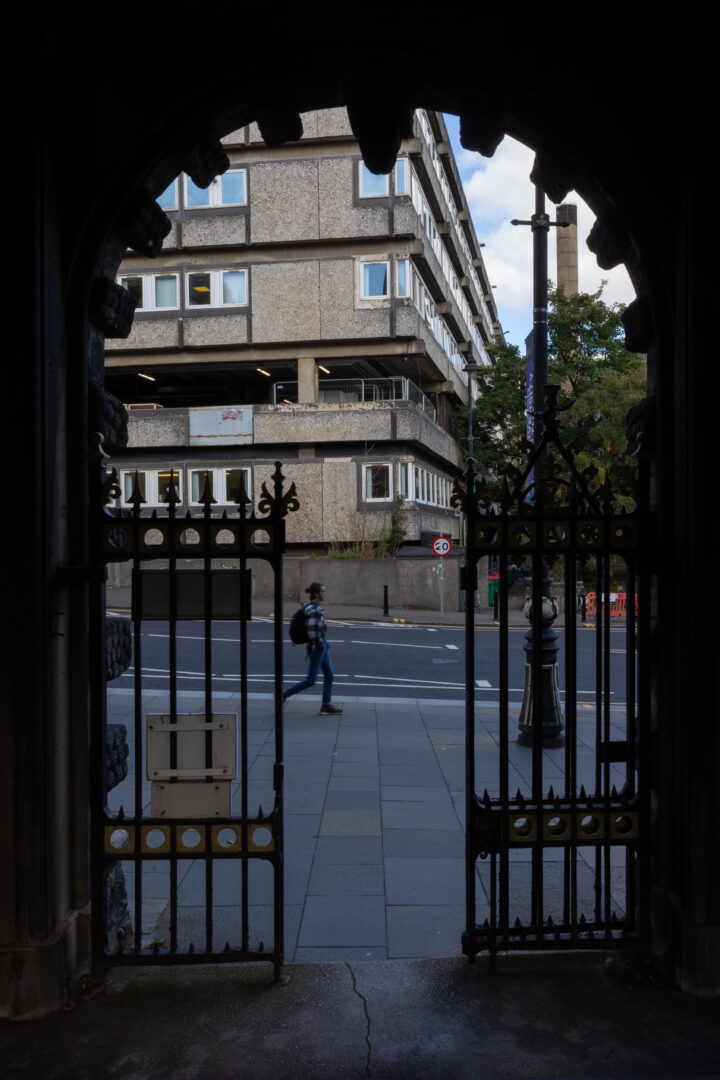 View through an ornate black iron gate of a pavement and street, with a pedestrian walking past. In the background, a modern, angular concrete building and some trees are visible under a partly cloudy sky.
