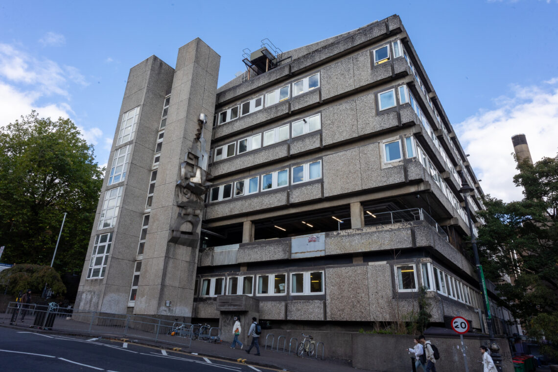 A large, grey, concrete Brutalist-style building with rectangular windows, angular features, and exterior staircases. People walk by on the pavement; trees and a blue sky with scattered clouds are visible in the background.