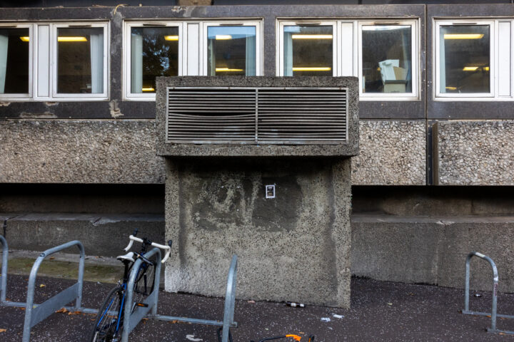 A concrete ventilation structure stands against a rough, pebbled building wall. Above it, rectangular windows line the building. A bicycle rack and a parked bike are visible in front, with scattered debris on the dark pavement.