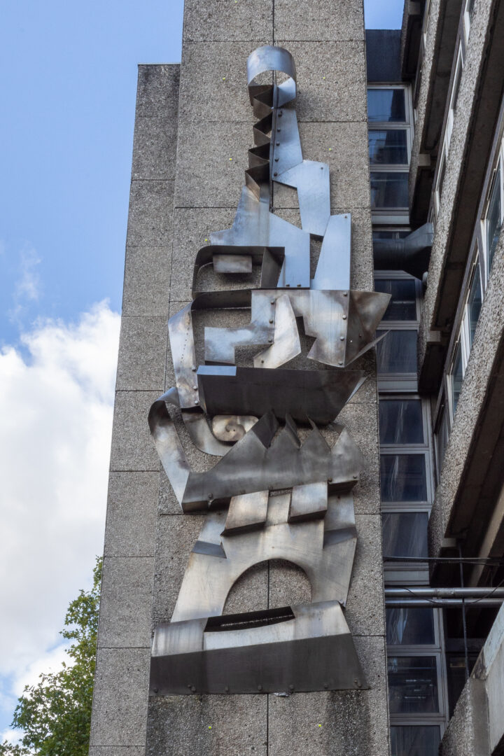A large, abstract metal sculpture is mounted vertically on a concrete building façade. The sculpture consists of curved and angular metallic shapes, creating a geometric design. Windows and a tree are visible nearby under a partly cloudy sky.