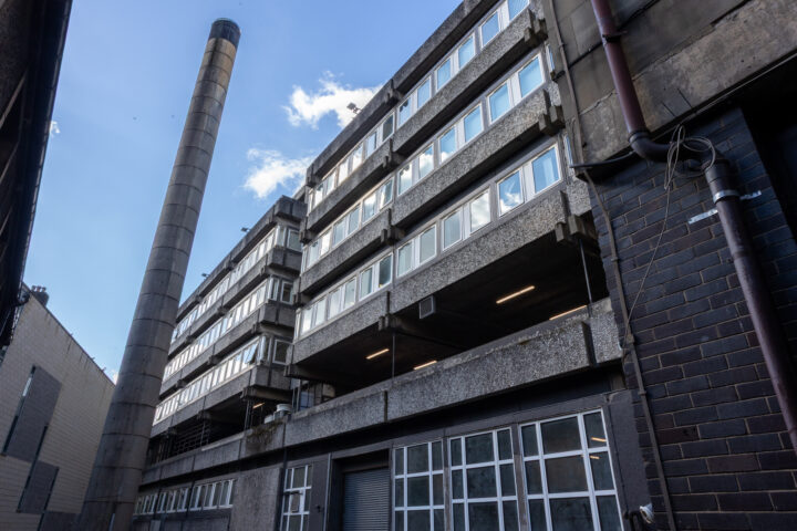 A brutalist-style building with exposed concrete, rows of rectangular windows, and a tall cylindrical chimney. The sky is blue with a few clouds visible. Pipes and industrial elements are seen along the building’s dark exterior wall.