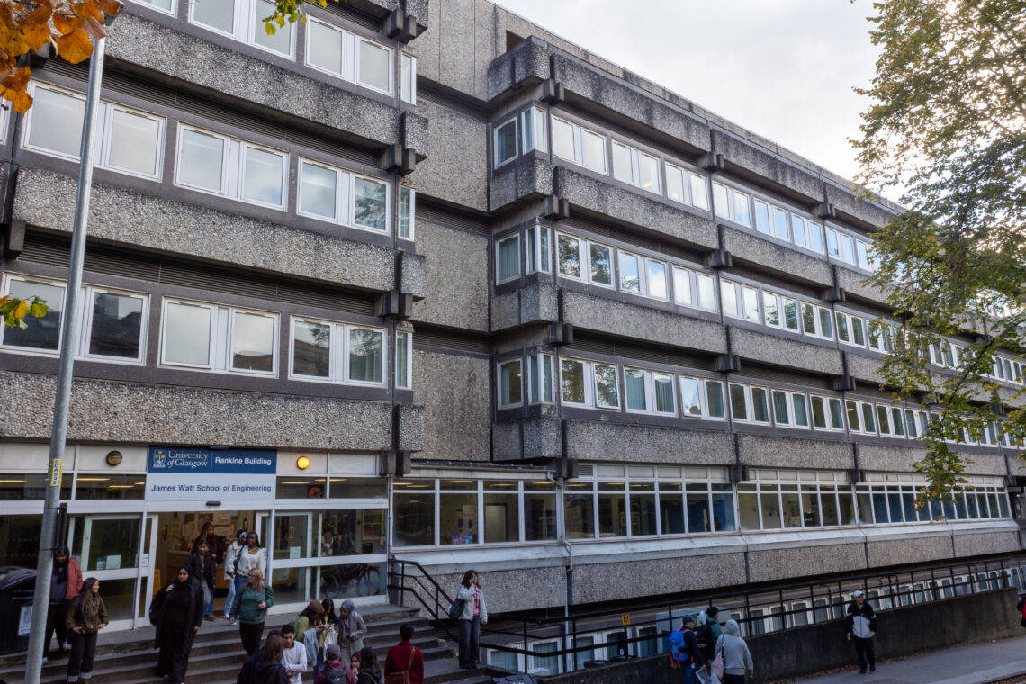 A large, grey concrete building with multiple rows of windows and a modernist design. People walk in and out of the entrance, where a blue sign reads James Watt School of Engineering. Trees frame the scene, and the sky is partly cloudy.
