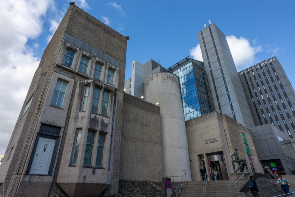 A modern university campus with concrete and glass buildings, including the University Library. The architecture features angular shapes, large windows, a cylindrical tower, and wide steps leading to entrances. People are walking near the buildings under a partly cloudy sky.