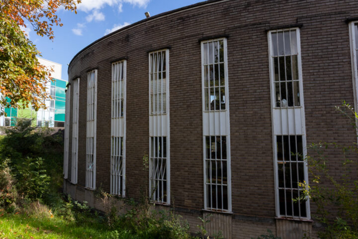 A curved brick building with tall, narrow windows, each covered by vertical metal bars. Greenery and overgrown plants are in the foreground, and a modern building with blue-green glass panels is visible in the background.