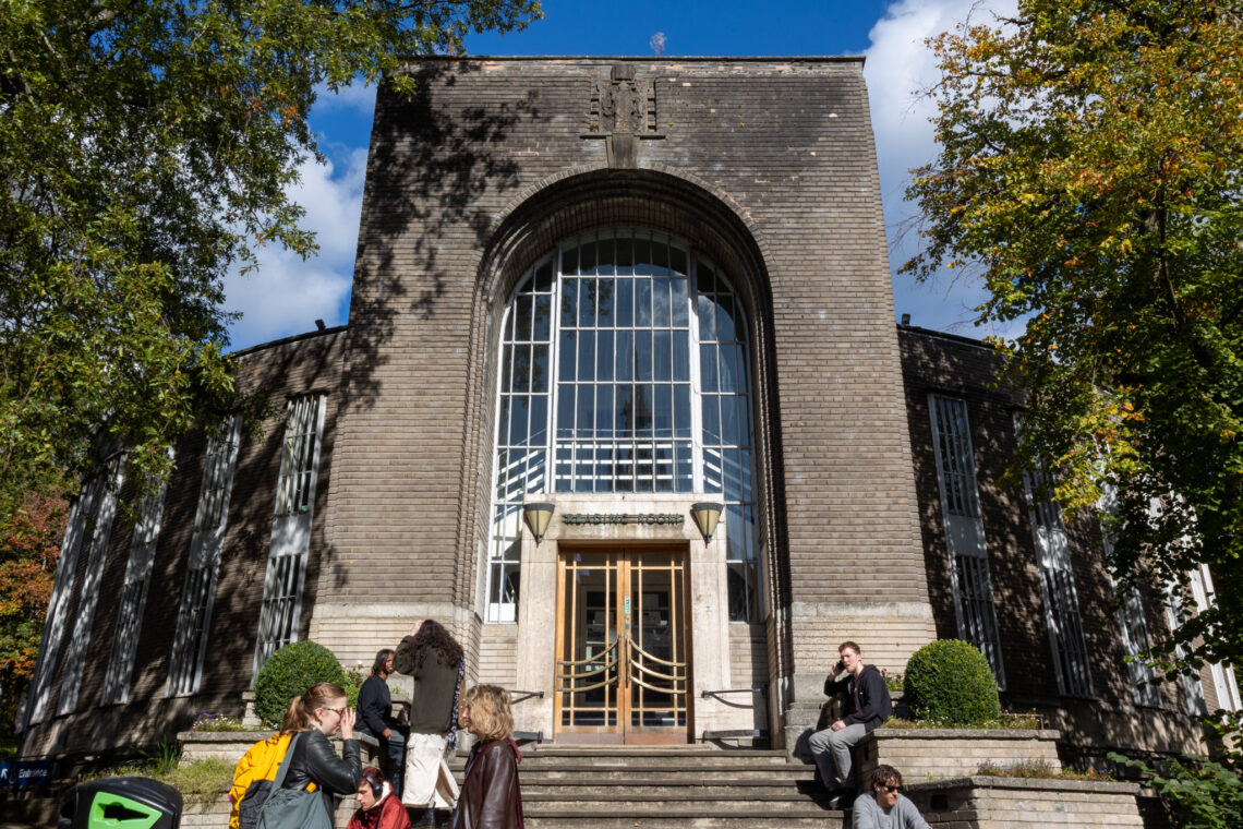 A large, brick building with a tall arched window and double glass doors at the entrance. Several people are gathered on the steps and walkway outside. Sunlight filters through trees flanking the building, and the sky is partly cloudy.