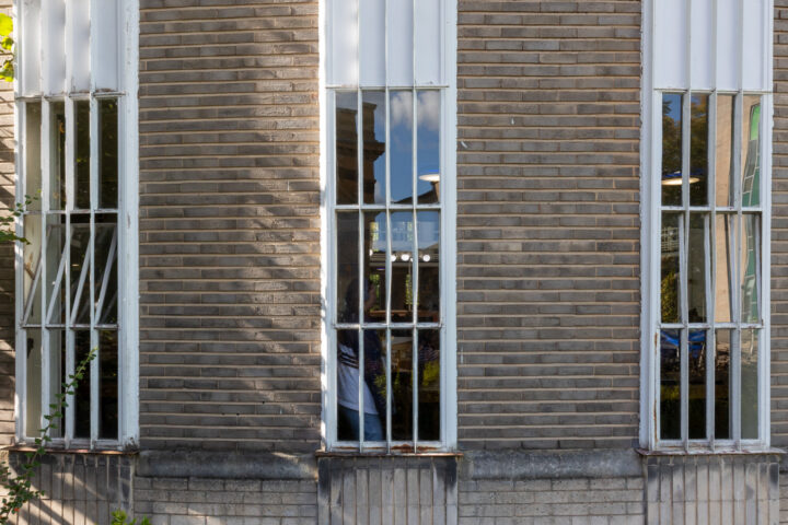 A close-up of a brick building façade with four tall, narrow windows. The windows have white frames and vertical bars. The glass reflects the blue sky, clouds, and parts of nearby buildings. Green foliage is partly visible on the left.