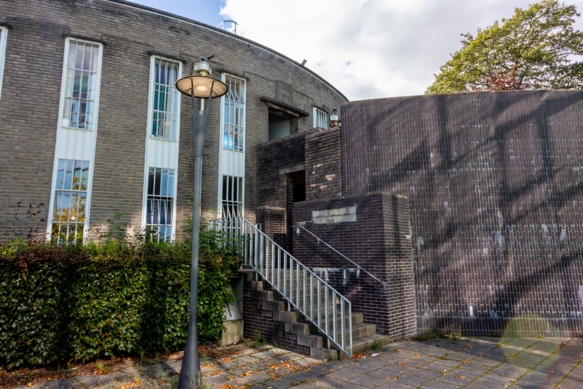 A curved brick building with tall narrow windows, a lamp-post, and an exterior staircase with a metal railing leading to a doorway. Bushes line the wall, and a tree with green and orange leaves is visible in the background under a partly cloudy sky.