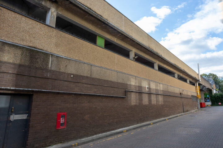 A long, multi-storey car park with brown brick and rough concrete walls. A red fire alarm box is mounted on the lower wall, and a green panel stands out on the upper level. The sky is partly cloudy, and the street below is empty.