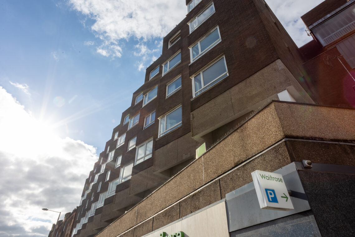 A stepped, modern brick building with many rectangular windows rises towards a partly cloudy blue sky. Sunlight creates a glare on the left. A white parking sign with a blue P and green Wichtrose is mounted on the lower right.