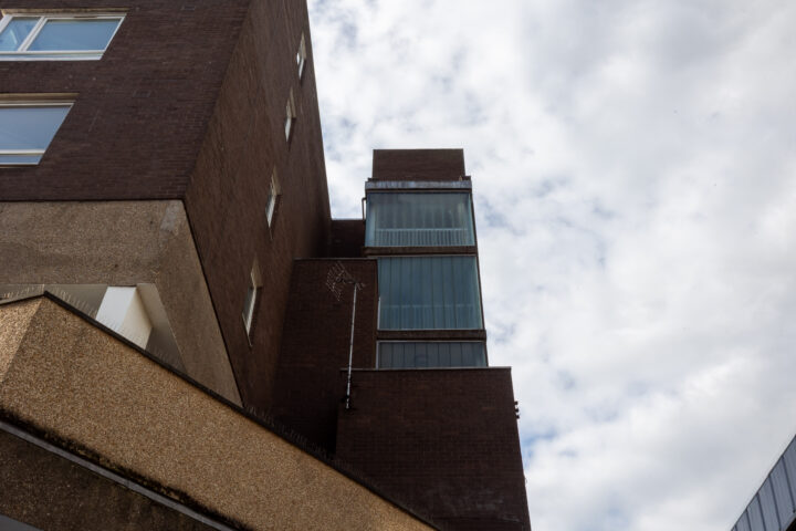 A modern, angular brown and beige building with large vertical glass windows rises towards a cloudy sky. The building has geometric shapes and sharp lines, contrasting with the soft, overcast clouds in the background.