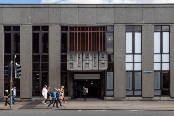 A concrete library building with tall narrow windows and a central entrance. Several people walk on the pavement in front, and a traffic light stands to the left. The sky is partly cloudy. Signs next to the entrance read “Library and Learning Centre.”.