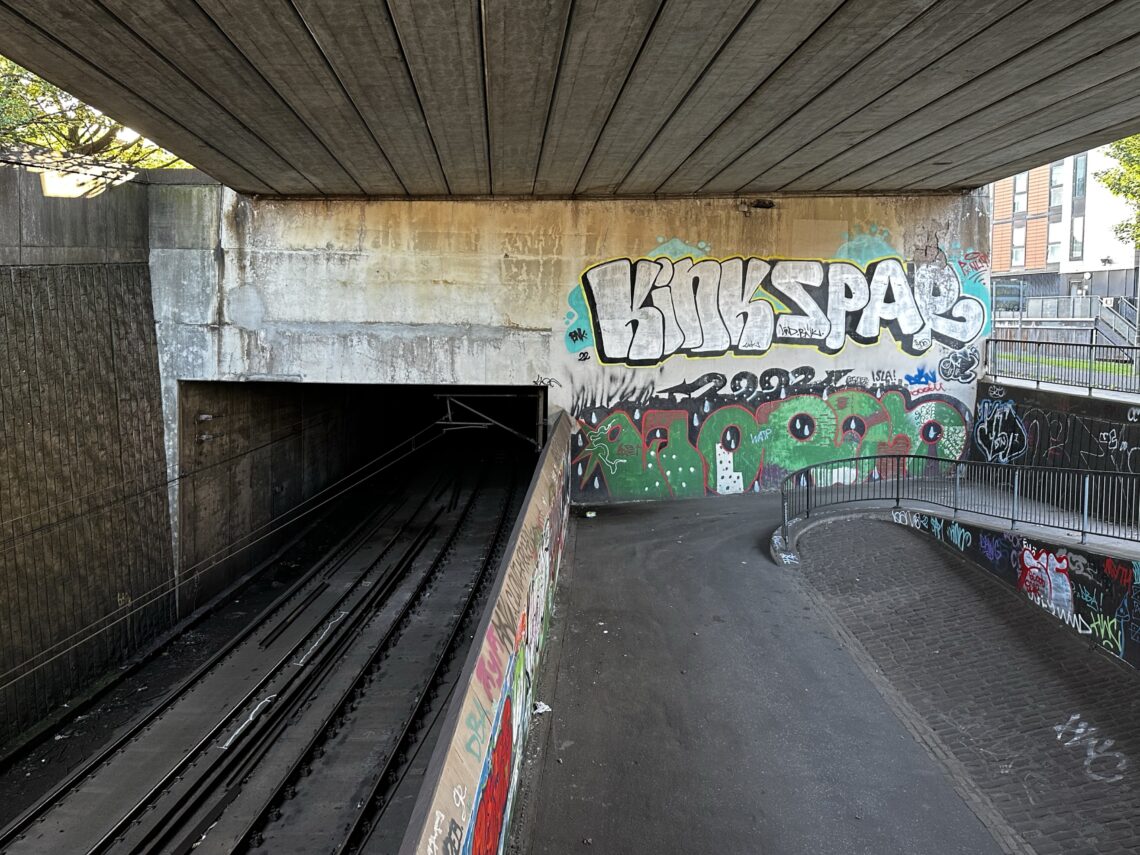 A railway tunnel entrance under a concrete bridge is covered in graffiti, including large white and silver text reading KINK SPAR. Railway tracks disappear into the dark tunnel, while a paved pedestrian path curves alongside fenced railings. Urban buildings are visible in the background.