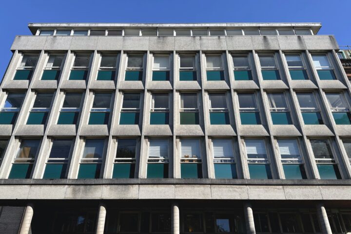 A five-storey modernist building with a grid of rectangular windows, teal and white panels, and concrete framing. The facade has strong vertical and horizontal lines, with a clear blue sky above. The ground level features columns and a stone exterior.
