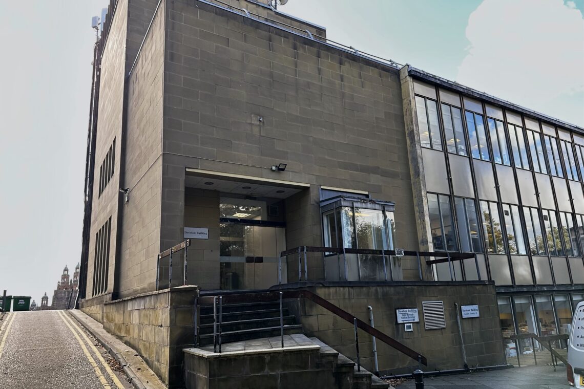 A modern, tan stone building with large glass windows sits beside a sloping street. The main entrance has steps, a handrail, a small canopy, and signs near the door. Reflections of trees and sky are visible in the windows.
