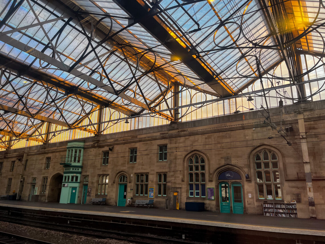 A historic railway station platform with an ornate glass roof letting in golden sunset light, casting warm reflections. The stone building has arched windows and doors, green trim, benches, and a bookcase. Railway tracks run in the foreground.