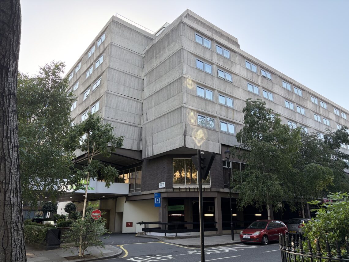 A large, grey concrete building with many windows stands on a street corner. Trees line the pavement. There’s a hotel entrance with a sign saying Hilton. Several parked cars and a no entry sign are visible near the entrance.
