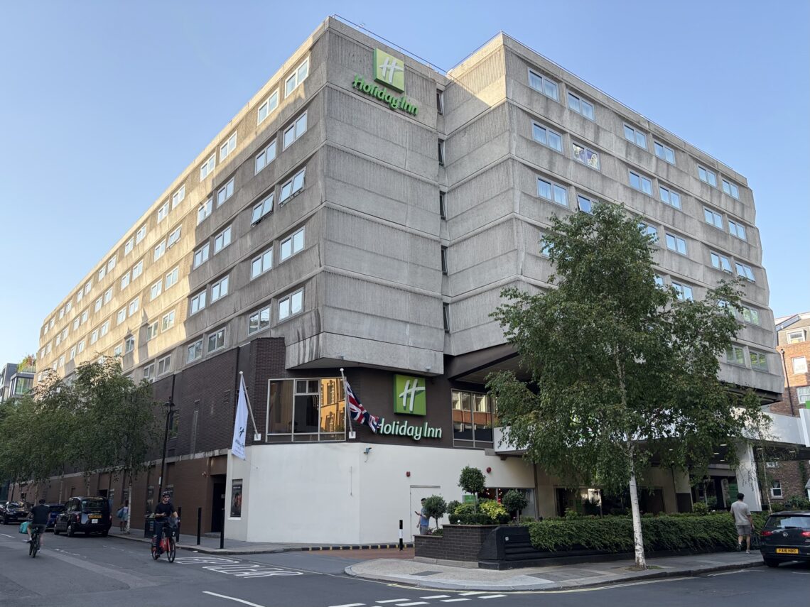 A large, modern Holiday Inn hotel with beige concrete exterior, green signage, and British flags at the entrance. Trees, a taxi, and people are visible on the street in front of the multi-storey building, which stands on a sunny corner in an urban area.