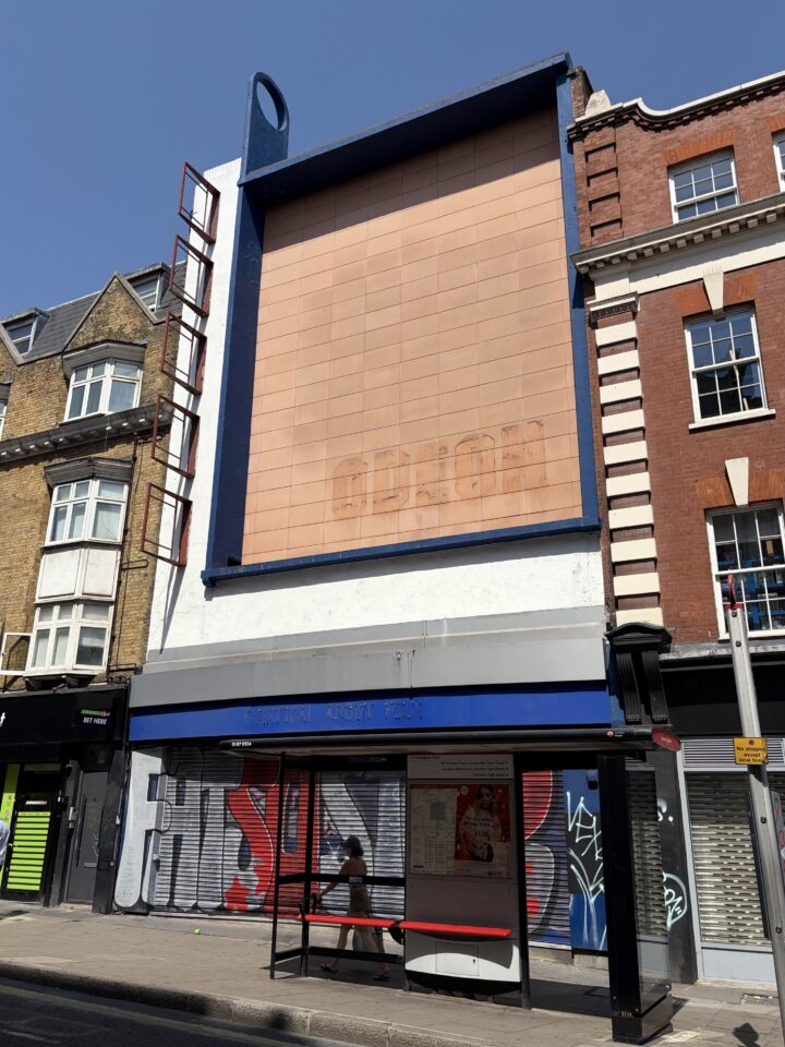 A large, faded peach-coloured billboard atop a closed shop displays faint, barely readable text ODEON. The shopfront below, with blue trim and metal shutters covered in graffiti, is flanked by brick buildings. A woman sits at a bus stop in front.