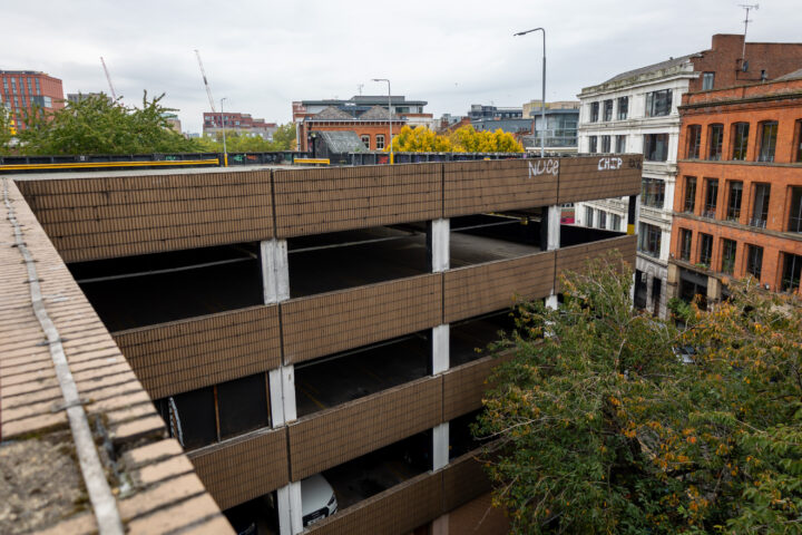A multi-storey brown car park with graffiti reading NUCE and CHIP on the top floor. Surrounding the car park are trees and neighbouring brick and white buildings. The sky is overcast, and cranes are visible in the distant background.