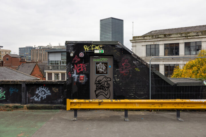 A rooftop scene shows a black door and wall covered in graffiti and tags, with a yellow metal barrier in front. Surrounding buildings, including a tall modern tower and older structures, are visible under a cloudy sky. Some trees with autumn leaves are also present.