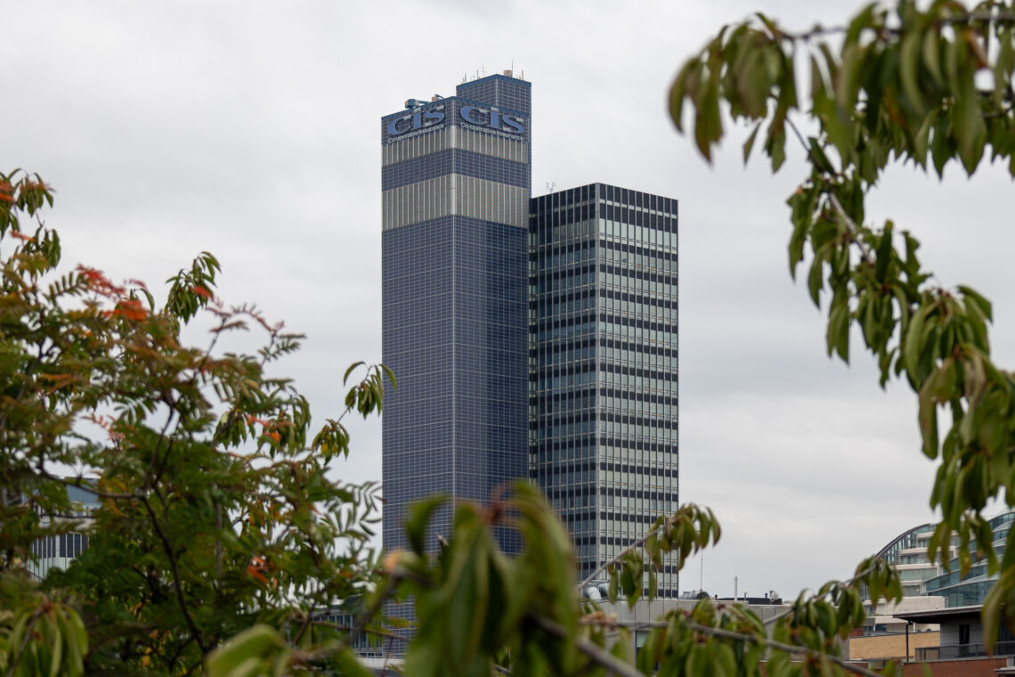 A tall, blue glass skyscraper with CIS CIS signage on top stands behind leafy green trees, under an overcast sky. The building features a grid pattern of windows and is accompanied by a slightly shorter, similarly styled tower.