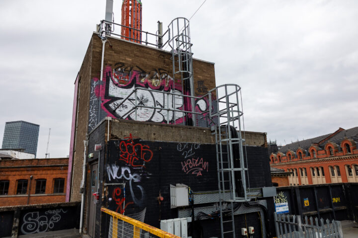 A brick building covered in colourful graffiti, with metal ladders and platforms attached. A tall orange structure stands on its roof. Surrounding buildings show old architecture and modern glass windows. The sky is overcast.