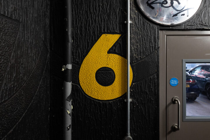 A large yellow number 6 is painted on a textured black wall. A metal pipe runs vertically in front of the number. To the right, there is a grey door with a window showing cars parked inside a garage. Above the door is a circular, partially obscured sign.