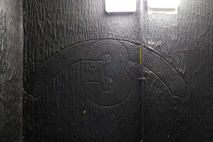 A rough black wall with a large, embossed number 3 centred. A raised arc curves over the top half of the 3. Two bright rectangular lights are above, with a conduit running vertically along the wall. The surface has a textured, uneven appearance.