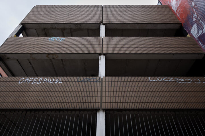 A low-angle view of a multi-storey concrete car park with horizontal brown tile panels, vertical beams, and some graffiti on the panels. The sky is overcast, and parts of a red mural are visible on the right edge.