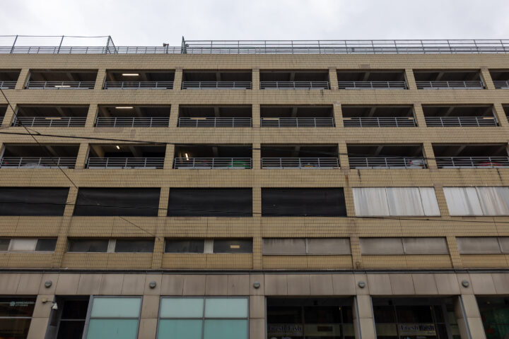 A beige multi-storey car park with horizontal metal railings on each level, dark and light window panels, and glass shopfronts on the ground floor. The sky is overcast, and the building appears vacant and functional.