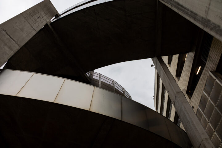 View looking up at intersecting concrete and metal structures of a car park against a cloudy sky. The architecture features curved and angled lines, creating a geometric pattern. Vertical and horizontal elements add depth and modern, industrial character.