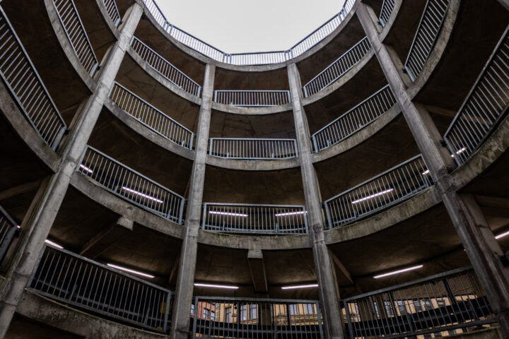 A circular concrete car park is shown from a low angle, looking up towards the open sky. Multiple levels feature metal railings and fluorescent lights, creating a pattern of repeating geometric shapes and lines.
