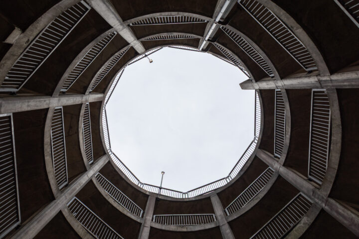 View looking upward from the centre of a circular, multi-level concrete car park. Spiral ramps and metal railings encircle an open skylight, revealing a cloudy sky above. Vertical support columns and geometric lines dominate the architectural structure.
