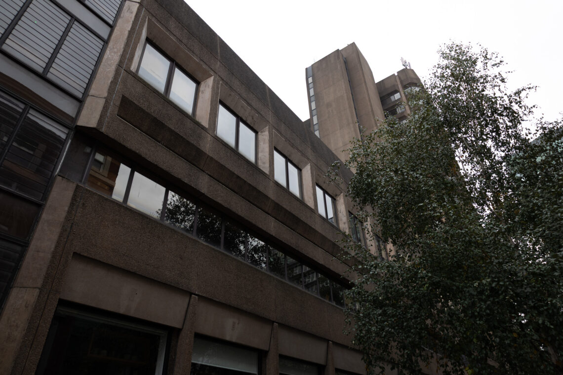 A low-angle view of a concrete Brutalist building with rectangular windows. A tree with green foliage stands in the foreground on the right, whilst the sky above is grey and overcast. The architecture features sharp lines and minimal decoration.
